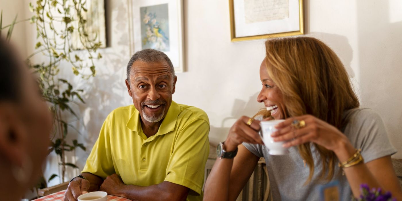Two people sit at a table, smiling and drinking coffee together in a warmly lit room with framed art on the wall.